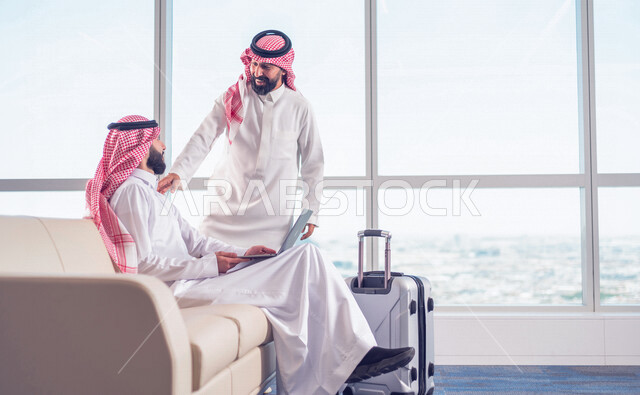 رجل اعمال عربي خليجي سعودي، يلتقي بزميل العمل اثناء الجلوس في قاعة الانتظار، انجاز العمل من خلال جهاز الحاسوب المحمول، العمل عن بعد، ايماءات وجه تدل على السعادة
