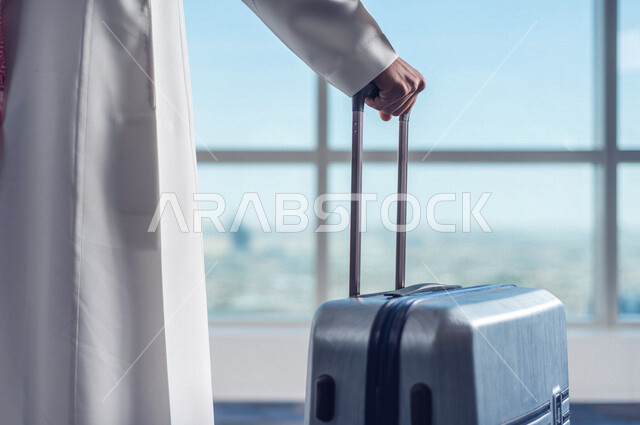 A Saudi Arabian Gulf businessman holding the travel bag and towing it, preparing to travel on a business trip, waiting lounge at the airport, tourism and travel, spending fun times