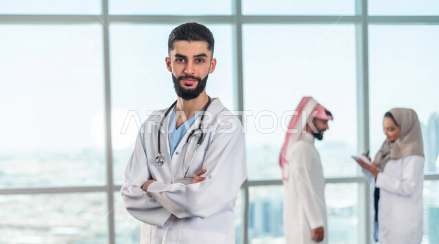 صورة مقربة لطبيب عربي خليجي سعودي ينظر الى الكاميرا وخلفية لفريق العمل، تبادل الاستشارات الطبية، تسجيل الملاحظات في دفتر الملاحظات، الالتزام باجراءات الوقاية من فايروس كورونا، مفهوم الطب والرعاية الصحية