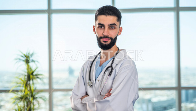 طبيب عربي خليجي سعودي ينظر إلى الكاميرا بإيماءات وجه ويدين متقاطعة، يقدم خدماته للمرضى بكل كفاءه، مفهوم الطب والرعاية الصحية