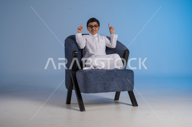 الجلوس والنظر إلى الكاميرا, بورتريه لفتى عربي خليجي سعودي مبتسم يرتدي الثوب التقليدي والنظارة الطبية يجلس على كنبه مريحة ويرفع اصابعه للأعلى, الإشارة إلى شيء ما بإيماءات السرور, خلفية زرقاء