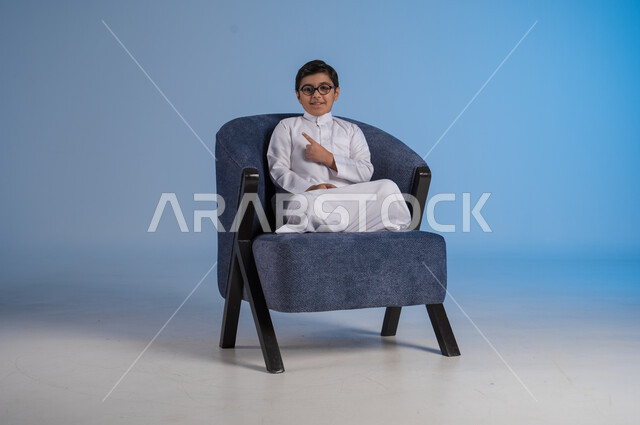 الجلوس والنظر إلى الكاميرا, بورتريه لفتى عربي خليجي سعودي مبتسم يرتدي الثوب التقليدي والنظارة الطبية يجلس على كنبه مريحة ويرفع اصبعه السبابة إلى جهة اليسار, الاشارة إلى شيء ما بإيماءات السرور, خلفية زرقاء
