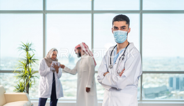 صورة مقربة لطبيب عربي خليجي سعودي ينظر الى الكاميرا وخلفية لفريق العمل، تبادل الاستشارات الطبية، تسجيل الملاحظات في دفتر الملاحظات، الالتزام باجراءات الوقاية من فايروس كورونا، مفهوم الطب والرعاية الصحية