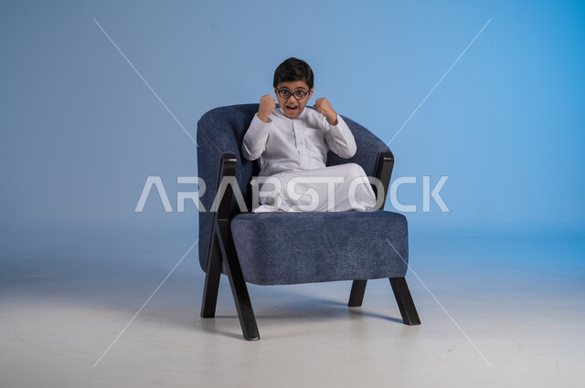 الانتصار والفوز والنجاح، مفهوم الانجاز وتحقيق الاهداف، بورتريه لفتى عربي خليجي سعودي يرتدي الثوب التقليدي والنظارة الطبية ينظر الى الكاميرا ويرفع يديه للاعلى بإيماءات الفرح والسرور, الشعور بالسعادة والقوة, خلفية بيضاء