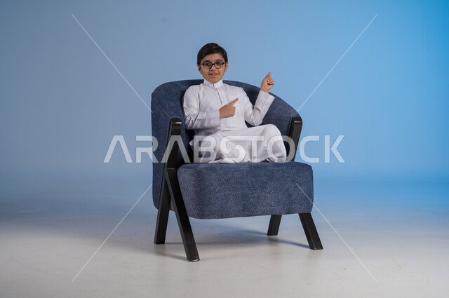 الاشارة إلى شيء ما وتحديد مكان معين, بورتريه لفتى عربي خليجي سعودي يرتدي الثوب التقليدي والنظارة الطبية يجلس على كنبه مريحة, النظر إلى الكاميرا بإيماءات السرور, خلفية رزقاء