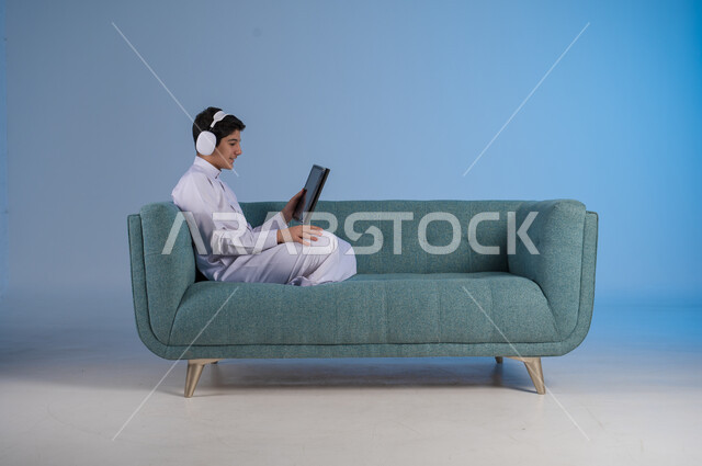 Listening to favorite music clips, entertainment and fun, using modern headphone technology, side portrait of a young Saudi Arabian Gulf man wearing traditional dress sitting on a comfortable sofa wearing audio headphones and holding a tablet in his hand, blue background