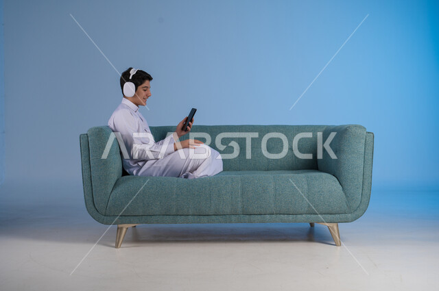 Having a good time listening to your favorite music tracks, using modern headphone technology, side portrait of a young Saudi Arabian Gulf man wearing traditional thobe sitting on a comfortable sofa wearing audio headphones and holding a mobile phone in his hand, blue background