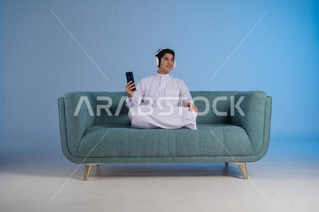 استخدام تقنية الهيدفون الحديثة, قضاء وقت ممتع بالاستماع إلى المقاطع الموسيقية المفضلة, بورتريه لشاب عربي خليجي سعودي يرتدي الثوب التقليدي يجلس على كنبه مريحة يضع سماعة الرأس الصوتية ويمسك بيده الهاتف المحمول, خلفية زرقاء