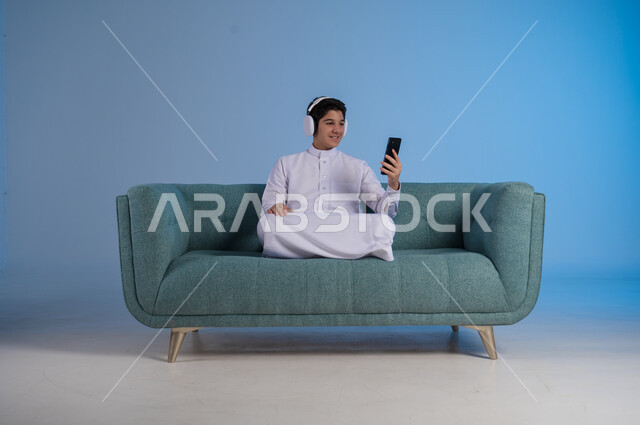 استخدام تقنية الهيدفون الحديثة, قضاء وقت ممتع بالاستماع إلى المقاطع الموسيقية المفضلة, بورتريه لشاب عربي خليجي سعودي يرتدي الثوب التقليدي يجلس على كنبه مريحة يضع سماعة الرأس الصوتية ويمسك بيده الهاتف المحمول, خلفية زرقاء