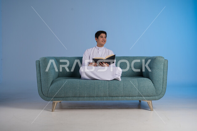 الأنشطة الترفيهية المفيدة, الاستمتاع بممارسة هواية المطالعة, بورتريه لشاب عربي خليجي سعودي مبتسم يرتدي الثوب التقليدي يجلس على كنبة مريحة ويمسك بيده الكتاب, النظر إلى شيء ما بإيماءات شرود الذهن, خلفية رزقاء