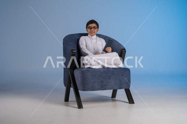 استخدام التكنولوجيا والتقنيات الحديثة والمتطورة، بورتريه لفتى عربي سعودي خليجي يرتدي الثوب التقليدي والنظارة الطبية يحمل بيده الهاتف المحمول وينظر إلى الكاميرا بسرور، خلفية زرقاء