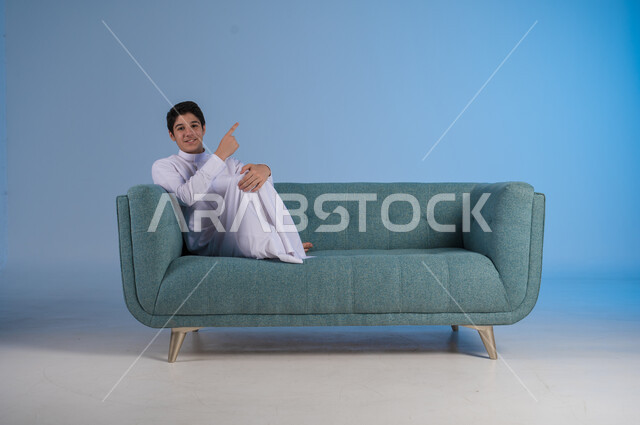 الاشارة إلى شيء ما وتحديد مكان معين, بورتريه لشاب عربي خليجي سعودي يرتدي الثوب التقليدي يجلس على كنبه مريحة, النظر إلى الكاميرا بإيماءات السرور, خلفية رزقاء