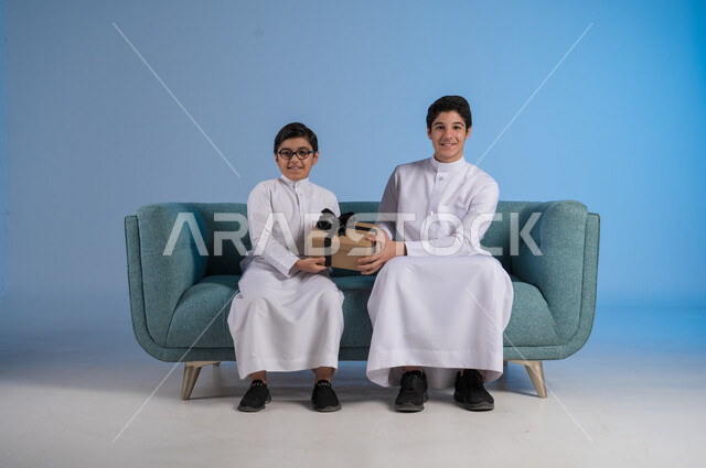 الألفة والمودة بين أفراد الأسرة السعودية, أعياد ومفاجآت ومناسبات سعيدة, مفهوم الإهداء والتقدير,  التعبير عن الحب والتقدير والاحترام, بورتريه لأخيين عربيين خليجيين سعوديين يرتديان الثوب التقليدي يحملان بيدهما علبة الهدية, خلفية زرقاء