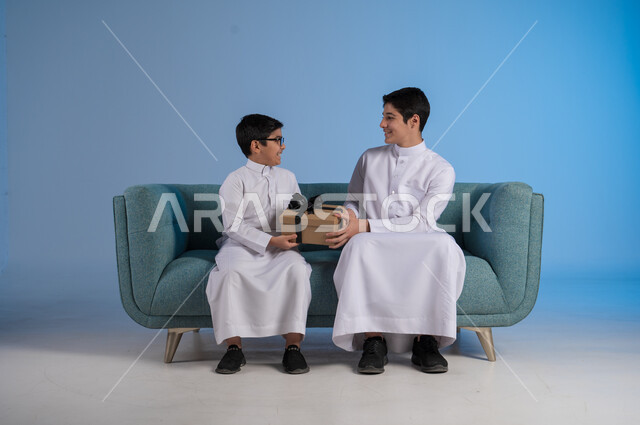 الألفة والمودة بين أفراد الأسرة السعودية, أعياد ومفاجآت ومناسبات سعيدة, مفهوم الإهداء والتقدير,  التعبير عن الحب والتقدير والاحترام, بورتريه لأخيين عربيين خليجيين سعوديين يرتديان الثوب التقليدي يحملان بيدهما علبة الهدية, خلفية زرقاء