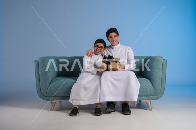 الألفة والمودة بين أفراد الأسرة السعودية, أعياد ومفاجآت ومناسبات سعيدة, مفهوم الإهداء والتقدير,  التعبير عن الحب والتقدير والاحترام, بورتريه لأخيين عربيين خليجيين سعوديين يرتديان الثوب التقليدي يحملان بيدهما علبة الهدية, خلفية زرقاء
