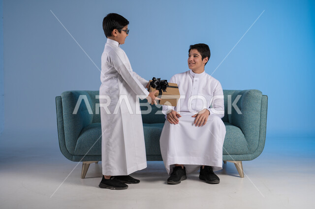 التعبير عن الحب والتقدير والاحترام, بورتريه لفتى عربي خليجي يرتدي الثوب التقليدي يقدم لأخيه علبة الهدية, أعياد ومفاجآت ومناسبات سعيدة, مفهوم الإهداء والتقدير, الألفة والمودة بين أفراد الأسرة السعودية, خلفية زرقاء