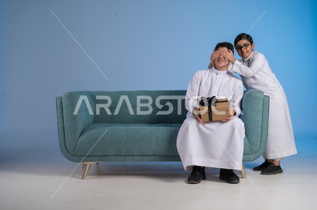 أعياد ومفاجآت ومناسبات سعيدة, مفهوم الإهداء والتقدير, الألفة والمودة بين أفراد الأسرة السعودية,  التعبير عن الحب والتقدير والاحترام, بورتريه لفتى عربي خليجي يرتدي الثوب التقليدي يقدم لأخيه علبة الهدية, خلفية زرقاء