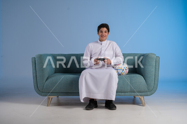 الأنشطة الترفيهية الممتعة, التقنيات الحديثة والتكنولوجيا, قضاء أوقات الفراغ باللعب بألعاب البلايستيشن, بورتريه لشاب عربي خليجي سعودي يرتدي الثوب التقليدي يمسك بيده عصا التحكم ويلعب بألعاب الفيديو بتفاعل واندماج, خلفية زرقاء