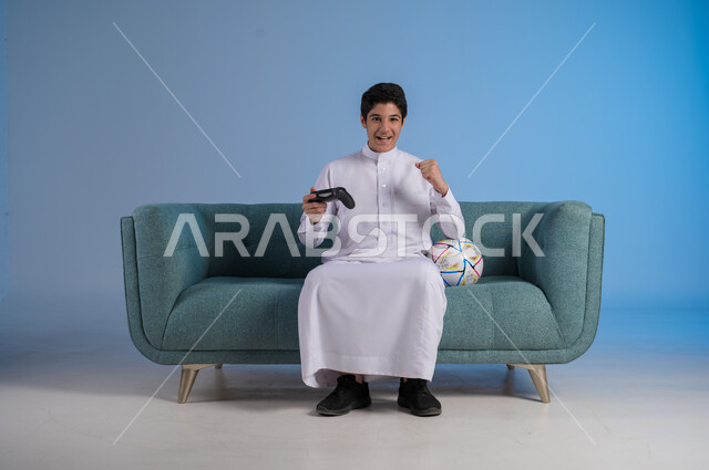 الأنشطة الترفيهية الممتعة, التقنيات الحديثة والتكنولوجيا, قضاء أوقات الفراغ باللعب بألعاب البلايستيشن, بورتريه لشاب عربي خليجي سعودي يرتدي الثوب التقليدي يمسك بيده عصا التحكم ويلعب بألعاب الفيديو بتفاعل واندماج, خلفية زرقاء