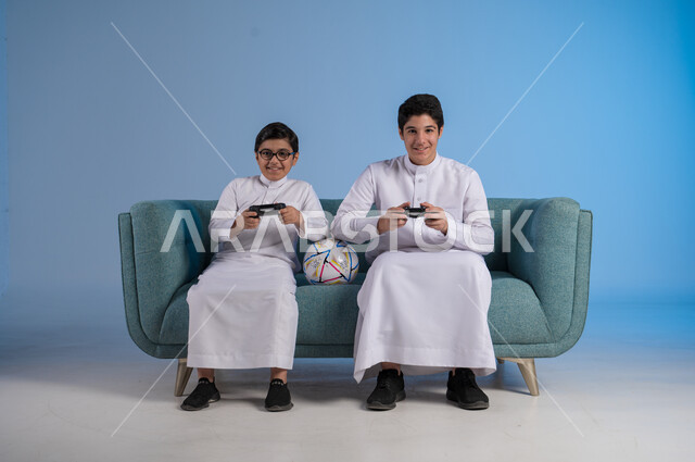 التسلية والترفيه وقضاء الوقت الممتع برفقة الأخوات, ألعاب البلايستيشن الترفيهية, بورتريه لصديقين عربيين خليجيين سعوديين يرتديان الثوب التقليدي يحملان عصا التحكم ويلعبان بألعاب الفيديو, المنافسة لتحقيق الانتصار, خلفية زرقاء