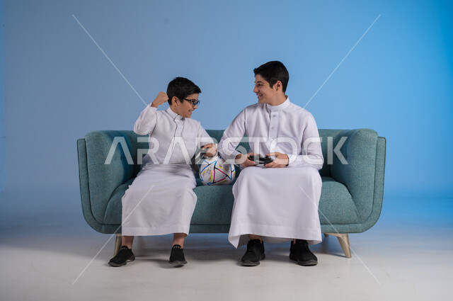 المنافسة لتحقيق الفوز والانتصار, ألعاب البلايستيشن الترفيهية, التسلية والترفيه وقضاء الوقت الممتع برفقة الأخوات, بورتريه لصديقين عربيين خليجيين سعوديين يرتديان الثوب التقليدي يحملان عصا التحكم ويلعبان بألعاب الفيديو, خلفية زرقاء