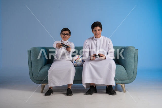 ألعاب البلايستيشن الترفيهية, التسلية والترفيه وقضاء الوقت الممتع برفقة الأخوات, بورتريه لصديقين عربيين خليجيين سعوديين يرتديان الثوب التقليدي يحملان عصا التحكم ويلعبان بألعاب الفيديو, المنافسة لتحقيق الانتصار, خلفية زرقاء