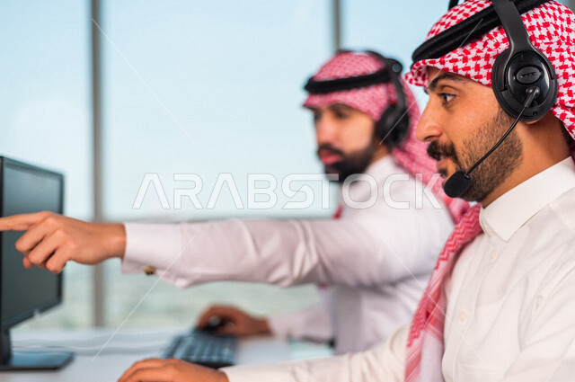 موظفان عربيان سعوديان خليجيان يعملان في خدمة العملاء في شركه يرتديان سماعة الاتصال, استعلامات واستفسارات, الاجابه على اسئلة العملاء, اجراء اتصال فيديو من خلال الحاسوب , عقد اجتماع عمل عبر الانترنت, العمل عن بعد , متابعة وتطوير العمل , مناقشة عمل , ارشاد و