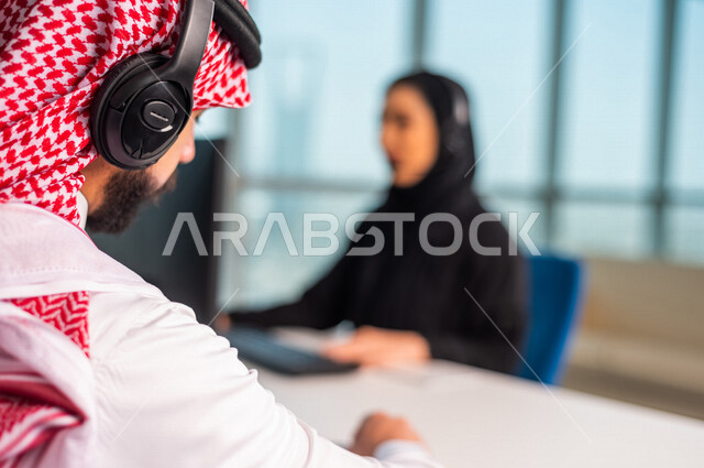 صوره مقربه  لخلفية رأس موظف  عربي سعودي خليجي في مقر العمل يرتدي  سماعة الاتصال والشماغ السعودي  . زملاء عمل في مركز الاستعلامات, اجراء اتصال فيديو من خلال الحاسوب , عقد اجتماع عمل عبر الانترنت , العمل عن بعد , متابعة وتطوير العمل