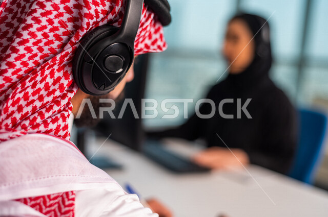 صوره مقربه  لخلفية رأس موظف  عربي سعودي خليجي في مقر العمل يرتدي  سماعة الاتصال والشماغ السعودي  . زملاء عمل في مركز الاستعلامات, اجراء اتصال فيديو من خلال الحاسوب , عقد اجتماع عمل عبر الانترنت , العمل عن بعد , متابعة وتطوير العمل