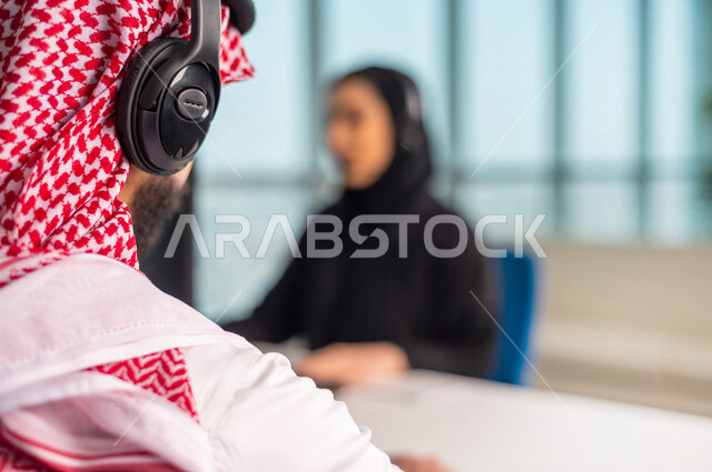 صوره مقربه  لخلفية رأس موظف  عربي سعودي خليجي في مقر العمل يرتدي  سماعة الاتصال والشماغ السعودي  . زملاء عمل في مركز الاستعلامات, اجراء اتصال فيديو من خلال الحاسوب , عقد اجتماع عمل عبر الانترنت , العمل عن بعد , متابعة وتطوير العمل