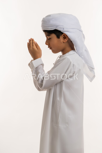 الاجتهاد في العبادة والتقرب الى الله، الرغبة في تحقيق الأمنيات، بورتريه من الجانب لولد عربي خليجي اماراتي يرتدي الكندورة والعمامة يرفع يديه للاعلى بايماءات الدعاء والتضرع، خلفية بيضاء