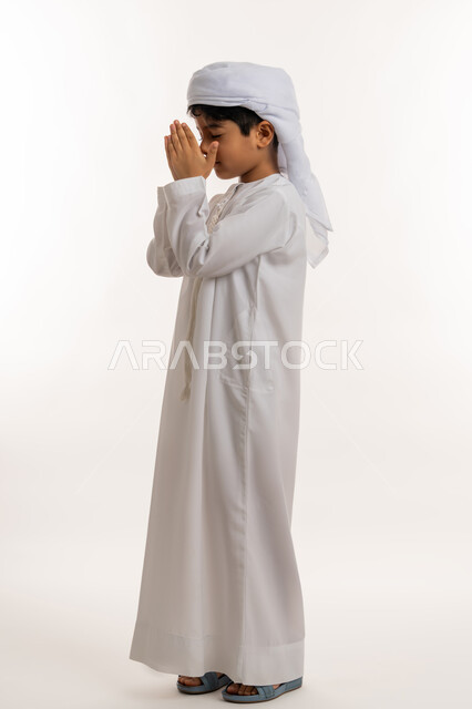 التقرب الى الله والاجتهاد في العبادة، الرغبة في تحقيق الأمنيات، بورتريه من الجانب لولد عربي خليجي اماراتي يرتدي الكندورة والعمامة يرفع يديه للاعلى بايماءات الدعاء والتضرع، صورة لكامل الجسم، خلفية بيضاء