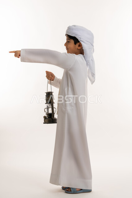 تحديد موقع شيء معين، الاستعداد لقدوم رمضان المبارك, زينة وديكورات شهر الخير، بورتريه من الجانب لولد عربي خليجي اماراتي يرتدي الكندورة والعمامة يحمل بيده فانوس مضيء يشير باصبع السبابة لمكان ما بإيماءات الفرح والسعادة، صورة لكامل الجسم، خلفية بيضاء
