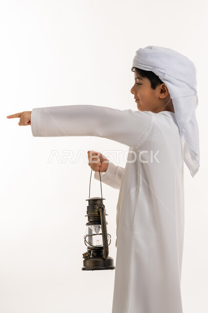 مصباح عربي معدني تقليدي، تحديد موقع شيء معين، الاستعداد لقدوم رمضان المبارك, زينة وديكورات شهر الخير، بورتريه من الجانب لولد عربي خليجي اماراتي يرتدي الكندورة والعمامة يحمل بيده فانوس مضيء يشير باصبع السبابة لمكان ما بإيماءات الفرح والسعادة، خلفية بيضاء