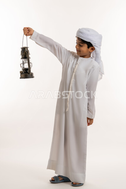 زينة وديكورات شهر الخير، الاحتفال بالاعياد والمناسبات الدينية، الاستعداد لقدوم رمضان المبارك, بورتريه من الجانب لولد عربي خليجي اماراتي يرتدي الكندورة والعمامة يحمل بيده فانوس مضيء ينظر لمكان ما بإيماءات الفرح والسعادة، صورة لكامل الجسم، خلفية بيضاء