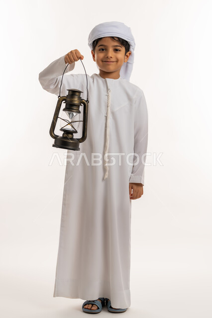 زينة وديكورات شهر الخير، الاحتفال بالاعياد والمناسبات الدينية، الاستعداد لقدوم رمضان المبارك, بورتريه لولد عربي خليجي اماراتي يرتدي الكندورة والعمامة يحمل بيده فانوس مضيء ينظر الى الكاميرا بإيماءات الفرح والسعادة، صورة لكامل الجسم، خلفية بيضاء