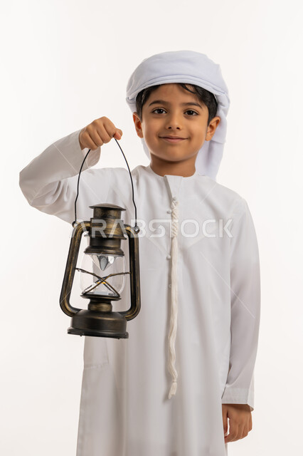 الاستعداد لقدوم رمضان المبارك, زينة وديكورات شهر الخير، الاحتفال بالاعياد والمناسبات الدينية، بورتريه من الجانب لولد عربي خليجي اماراتي يرتدي الكندورة والعمامة يحمل بيده فانوس مضيء ينظر الى الكاميرا بإيماءات الفرح والسعادة، خلفية بيضاء