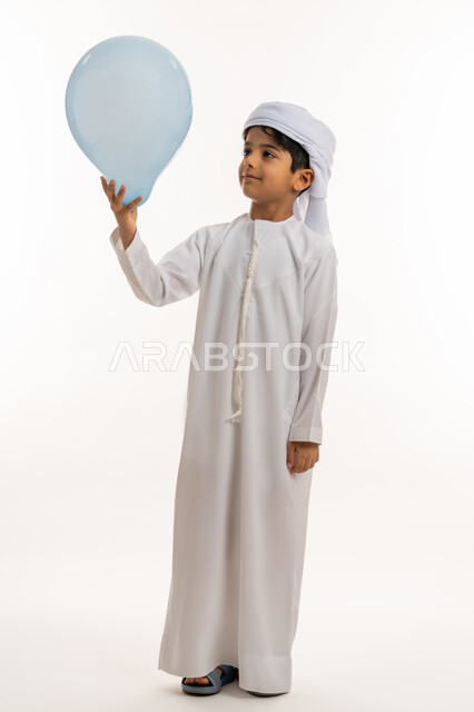 الاحتفال بالأعياد والمناسبات الدينية، فرحة وبهجة الاولاد في العيد، تعابيرالسرور والسعادة، بورتريه لولد عربي خليجي اماراتي يرتدي الكندورة والعمامة تحمل بيدها بالون ازرق، صورة لكامل الجسم، خلفية بيضاء