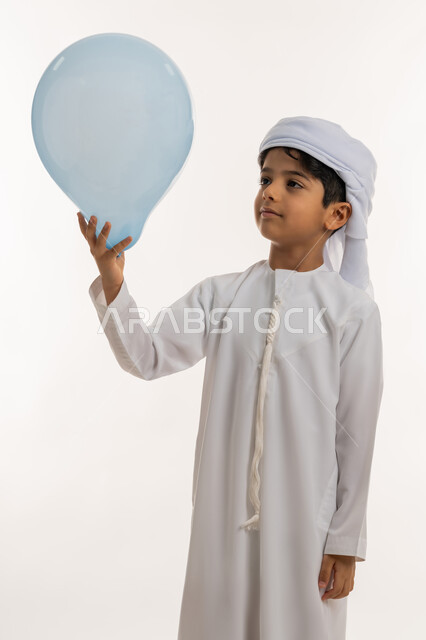 Gestures of joy and happiness, celebrating religious holidays and occasions, joy and happiness of children on Eid, portrait of an Arab Gulf Emirati boy wearing a kandura and turban holding a blue balloon in his hand, white background
