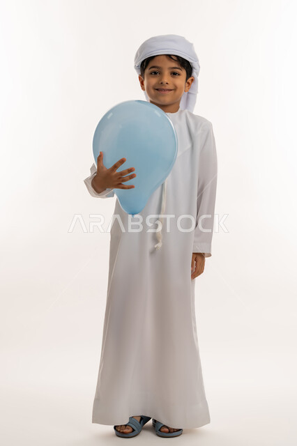 فرحة وبهجة الاولاد في العيد، الاحتفال بالأعياد والمناسبات الدينية، تعابيرالسرور والسعادة، بورتريه لولد عربي خليجي اماراتي يرتدي الكندورة والعمامة تحمل بيدها بالون ازرق، صورة لكامل الجسم، خلفية بيضاء