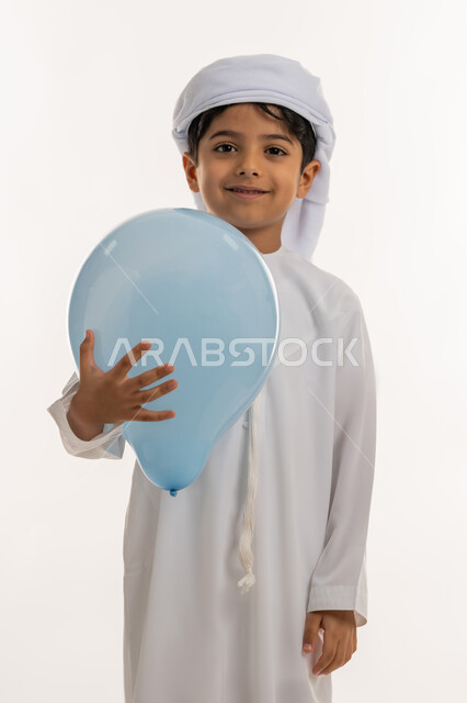 Expressions of joy and happiness, celebrating religious holidays and occasions, joy and happiness of children on Eid, portrait of an Arab Gulf Emirati boy wearing a kandura and turban holding a blue balloon in his hand, white background
