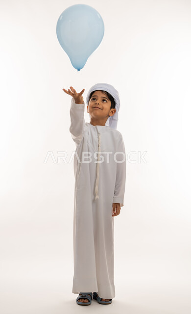 فرحة وبهجة الاولاد في العيد، الاحتفال بالأعياد والمناسبات الدينية، تعابيرالسرور والسعادة، بورتريه لولد عربي خليجي اماراتي يرتدي الكندورة والعمامة تحمل بيدها بالون ازرق، صورة لكامل الجسم، خلفية بيضاء
