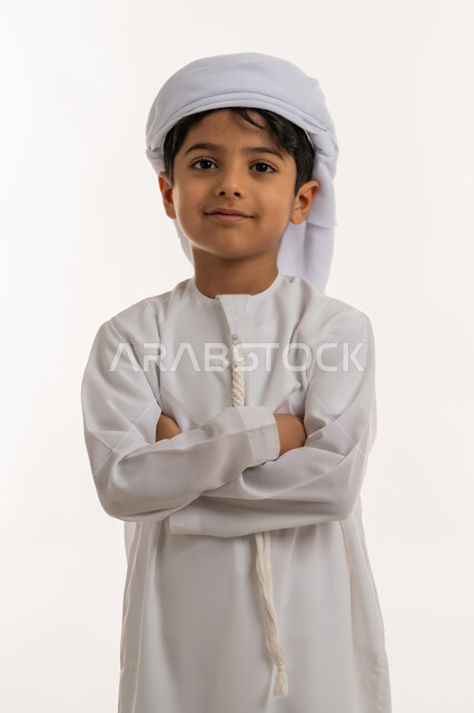 الاهتمام بالأناقة والمظهر الخارجي، تعابير السرور والسعادة، بورتريه لولد عربي خليجي اماراتي مبتسم يرتدي االعمامة والكندورة يقف بايدي متقاطعة ينظر الى الكاميرا بإيماءات الثقة بالنفس، خلفية بيضاء