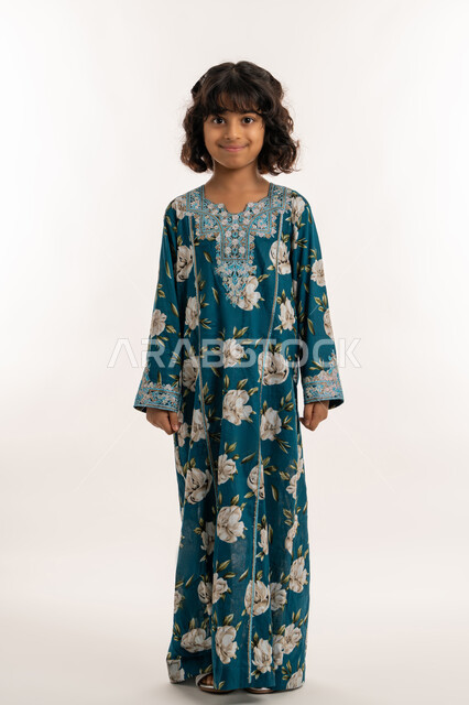 النظر الى الكاميرا بايماءات الثقة بالنفس، حب التزين وارتداء الملابس الجديدة، مفهوم الاناقة والنعومة، الاهتمام بالمظهر الخارجي، بورتريه لفتاة عربية خليجية اماراتية ترتدي فستان العيد، بنت سعودية انيقة تقف باستقامة، صورة لكامل الجسم، خلفية بيضاء