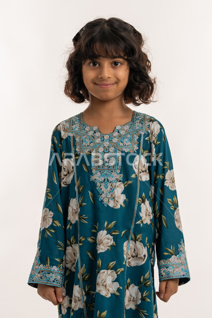 النظر الى الكاميرا بايماءات الثقة بالنفس، حب التزين وارتداء الملابس الجديدة، مفهوم الاناقة والنعومة، الاهتمام بالمظهر الخارجي، بورتريه لفتاة عربية خليجية اماراتية ترتدي فستان العيد، بنت سعودية انيقة تقف باستقامة، خلفية بيضاء