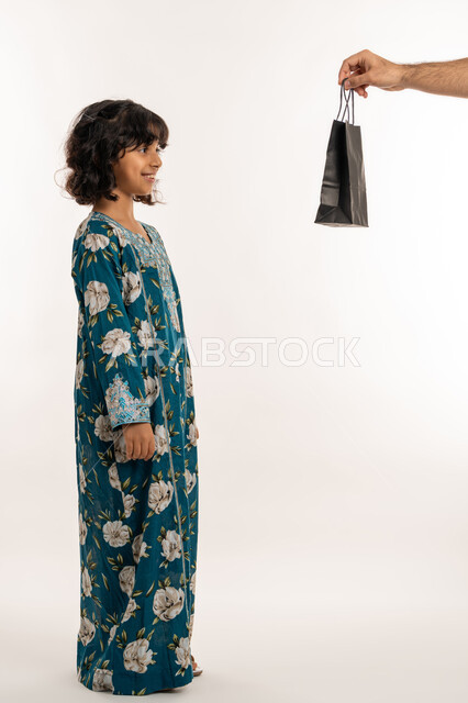 ادخال السرور الى قلب الابناء، فرحة وبهجة الاولاد في العيد، الاحتفال بالأعياد والمناسبات الدينية، تعابيرالفرح والسعادة، بورتريه من الجانب لفتاة عربية خليجية اماراتية ترتدي فستان تقوم بتلقي الهدايا من والدها، بنت سعودية انيقة، صورة لكامل الجسم، خلفية بيضاء