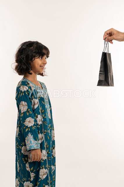 فرحة وبهجة الاولاد في العيد، ادخال السرور الى قلب الابناء، الاحتفال بالأعياد والمناسبات الدينية، تعابيرالفرح والسعادة، بورتريه من الجانب لفتاة عربية خليجية اماراتية ترتدي فستان تقوم بتلقي الهدايا من والدها، بنت سعودية انيقة، خلفية بيضاء
