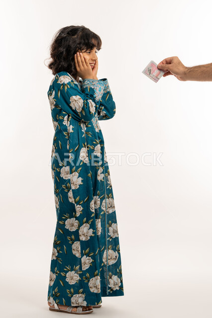 قضاء أوقات ممتعة مع العائلة، ايماءات الفرحة و البهجة، المناسبات والاعياد الدينية، بورتريه من الجانب لفتاة عربية خليجية اماراتية ترتدي فستان تقوم بتلقي العيدية من والدها، الاحتفال بقدوم العيد،  بنت سعودية تضع يديها على وجهها بتعابير الدهشة، صورة لكامل الجسم، خلفية بيضاء