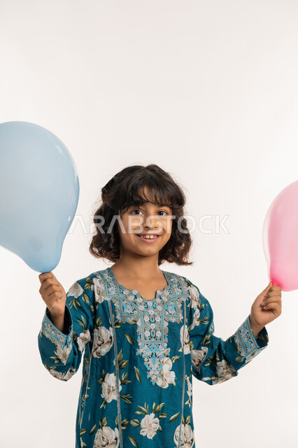 الاحتفال بالأعياد والمناسبات الدينية، النظر الى الكاميرا بتعابيرالسرور والسعادة، فرحة وبهجة الاولاد في العيد، بورتريه مقرب لفتاة عربية خليجية اماراتية ترتدي جلابية بناتية تحمل بيدها بوالين ملونة، بنت سعودية تقف بايماءات الفرح، خلفية بيضاء