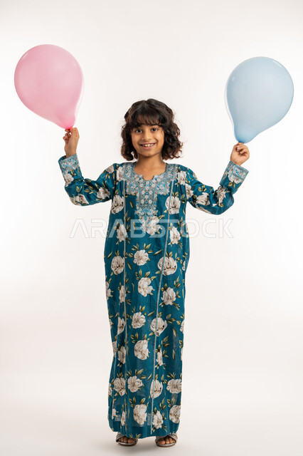 فرحة وبهجة الاولاد في العيد، النظر الى الكاميرا بتعابيرالسرور والسعادة، الاحتفال بالأعياد والمناسبات الدينية، بورتريه لفتاة عربية خليجية اماراتية ترتدي جلابية بناتية تحمل بيدها بوالين ملونة، بنت سعودية تقف بايماءات الفرح، صورة لكامل الجسم، خلفية بيضاء